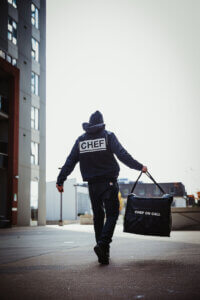 A man walks down the street carrying a bag labeled "CHEF on Call Delivery," suggesting he is a fast food delivery professional.