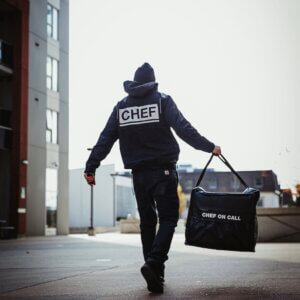 A man walks down the street carrying a bag labeled "CHEF on Call Delivery," suggesting he is a fast food delivery professional.