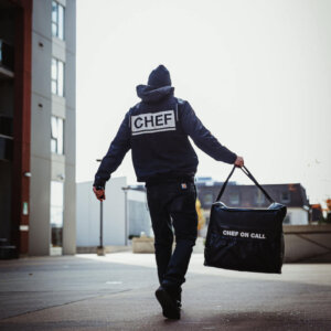 A man walks down the street carrying a bag labeled "CHEF on Call Delivery," suggesting he is a fast food delivery professional.